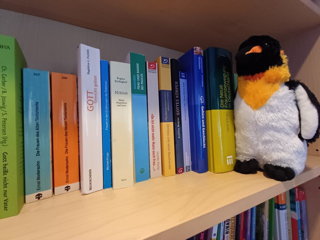 Junia in einem Teil ihrer Spezialbibliothek - Pinguin sitzt vor einigen Büchern, u.a. Christus und seine Geschwister, Gott Gewicht geben, Frau Mann und Paulus, Gehirn und Geschlecht, Da ist nicht Mann noch Frau..., Gottes Körper,  Gehirn und Geschlecht, Die Frauen im Alten Testament, Die Frauen im Neuen Testament, Frauen und Erzählungen der Rabbinen, Frauen Männer, Engel,
