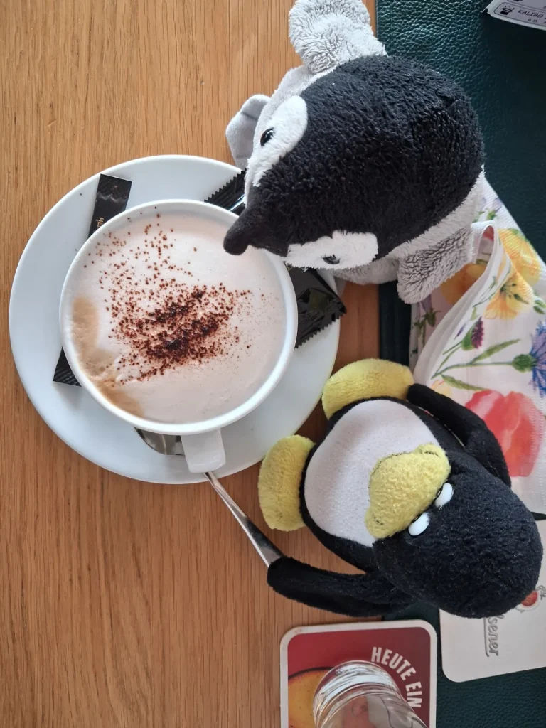 Neville und Mütux sitzen bei einer Tasse Capuchino zusammen.Mütux hebt grad den Löffeln zum Umrühren an.