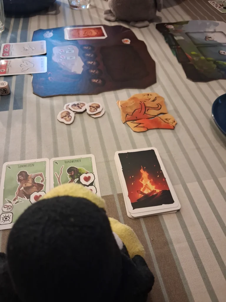 Mütux und Neville spielen Paleo. Vor ihnen sind die Karten ausgelegt, 2 Teile der Mammutzeichnung haben sie bereits gewonnen.
