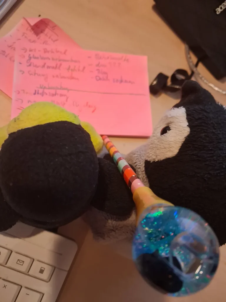 Neville und Mütux schreiben gemeinsam mit einem Multifarbenbuntstift auf ein rotes PostIT. Oben am Stift ist eine kleine Kugel mit einem Pinguin drin.