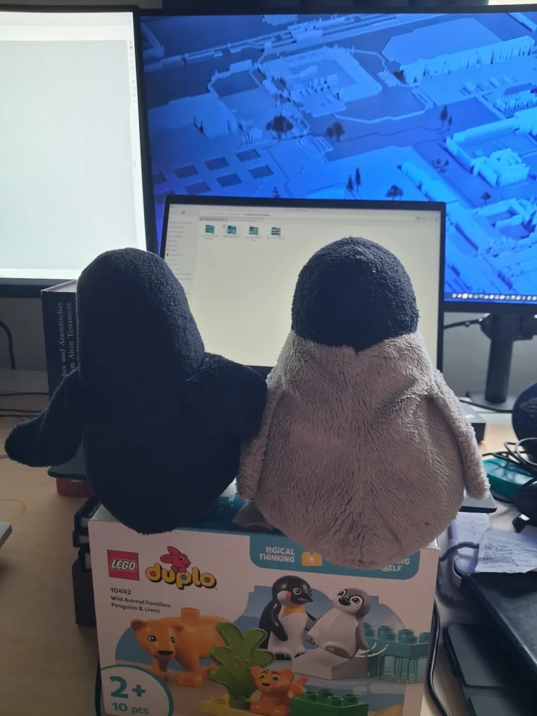 Mütux und Neville sitzen auf einem Pinguin DuploKarton und schauen auf den Laptop, dahinter sind 2 weitere Bildschirme.