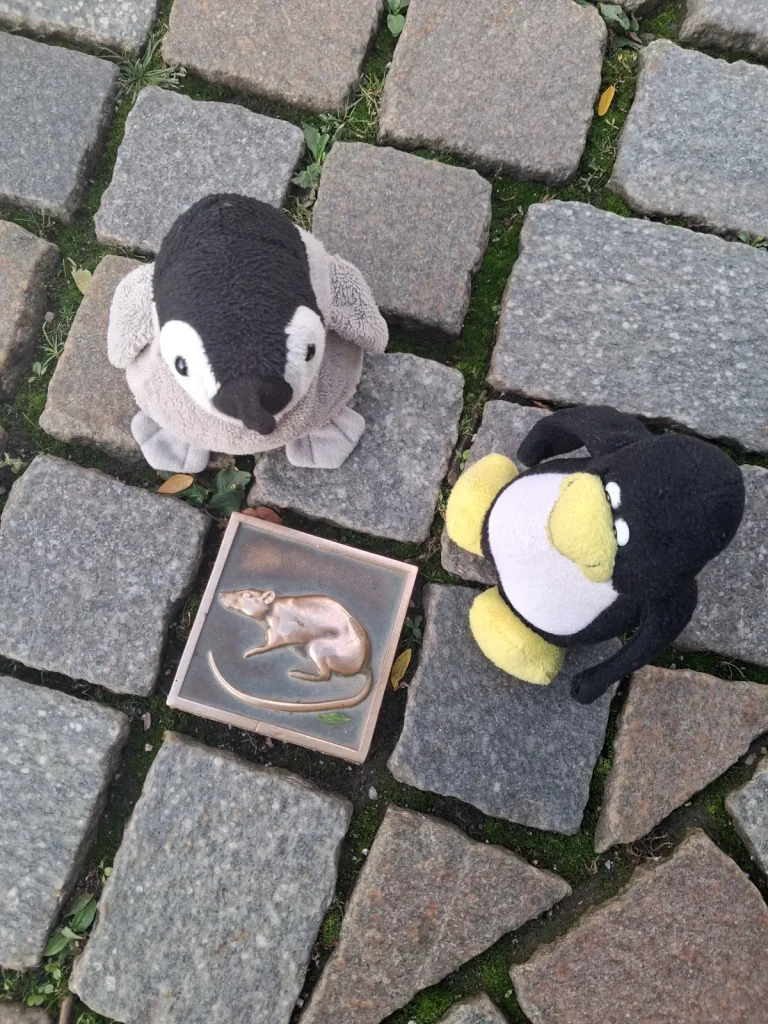 Mütux und Neville sitzen auf einem gepflasterten Boden und schauen auf einen goldenen Stein mit einer Ratte.