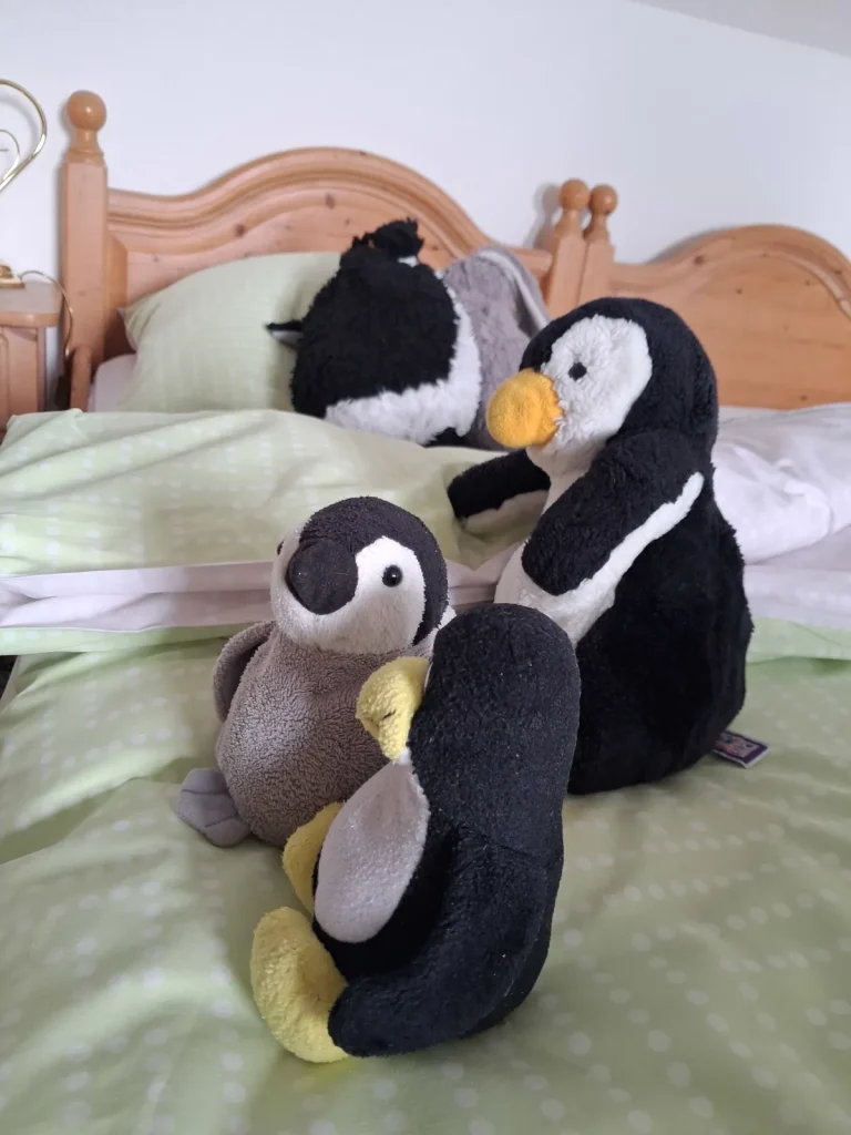 Neville und Mütux schauen in Richtung Kamera, Pingu sitzt dahin. Alle drei auf einer grünen Bettdecke. Im Hintergrund ruehen sich Junior und Klopfer von der anstrengenden Reise aus.