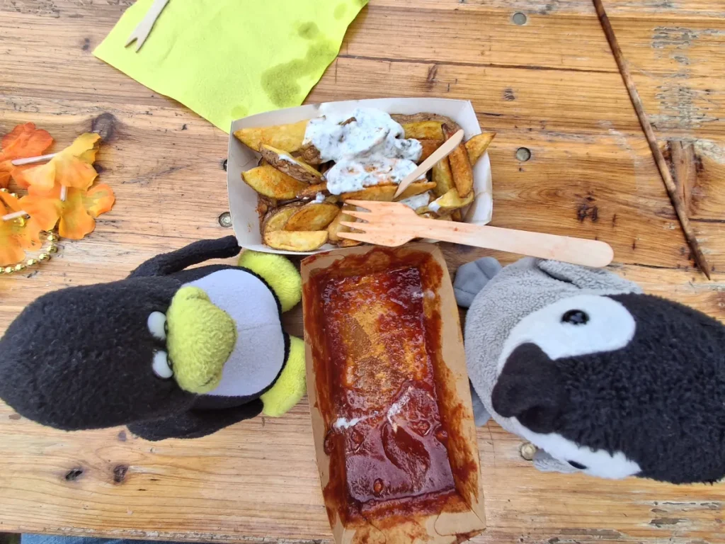 Auf einem rustikalen Holztisch stehen zwei Mütux und Neville neben einer Mahlzeit. In der Mitte liegen knusprige Kartoffelspalten mit einem hellen Kräuterdip sowie ein längliches Gericht, das mit roter Soße bedeckt ist. Zwei kleine Holzgabeln liegen bei den Speisen. Links am Bildrand sind orangefarbene Blüten zu sehen, oben eine grüne Serviette. Die Aufnahme wirkt wie eine humorvolle Szene, in der die beiden Pinguine gemeinsam am Tisch essen.