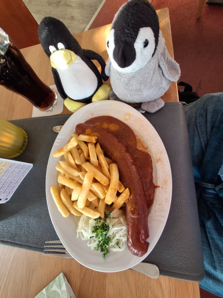 Auf dem Bild sitzen Mütux und Neville gemeinsam an einem Restauranttisch und haben ihr Essen vor sich. Auf dem großen Teller liegt eine Portion Currywurst mit roter Soße und Currypulver, dazu Pommes frites sowie etwas Krautsalat mit Kräutern. Links daneben steht ein großes Glas mit einem dunklen Getränk.
Mütux und Neville sitzen direkt hinter dem Teller und schauen auf die Mahlzeit, als würden sie gespannt darauf warten, dass es endlich losgeht. Durch die Perspektive von oben stehen die beiden Pinguine und das üppige Essen klar im Mittelpunkt der Szene.