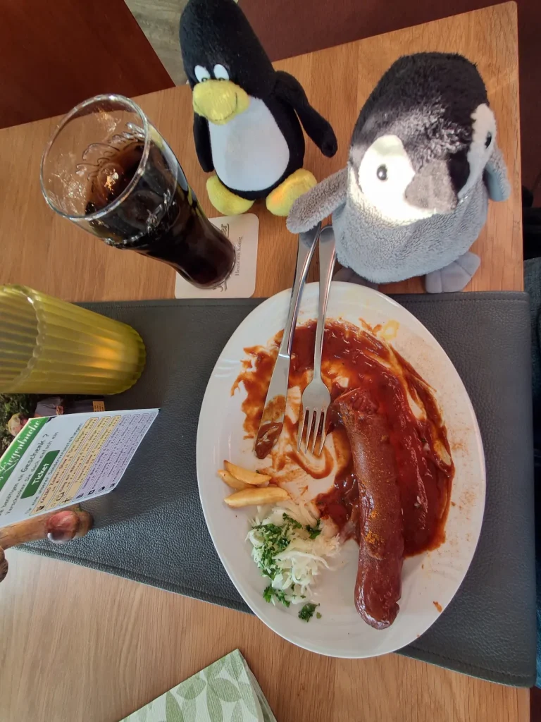 Auf dem Bild sitzen **Mütux und Neville** wieder gemeinsam am Restauranttisch. Vor ihnen steht der Teller mit der **Currywurst**, von der bereits ein großer Teil gegessen wurde. Nur noch etwas Wurst, wenige Pommes und ein kleiner Rest Krautsalat sind übrig; Messer und Gabel liegen mitten auf dem Teller.
Links daneben steht weiterhin das große Glas mit dem dunklen Getränk. **Mütux und Neville** sitzen dicht hinter dem Teller und wirken, als würden sie zufrieden auf die fast vollständig verspeiste Mahlzeit schauen. Das Bild vermittelt den Eindruck, dass die beiden ihr Essen ausgiebig genossen haben.