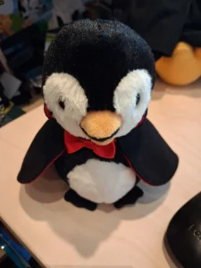 **Bildbeschreibung: Nibbie, der Vampirpinguin**
Auf dem Bild ist Nibbie zu sehen, ein kleiner, rundlicher Plüschpinguin mit einem niedlichen Vampirkostüm. Sein Körper ist überwiegend schwarz, mit einem flauschigen weißen Bauch und großen weißen Gesichtspartien. Er hat einen orangefarbenen Schnabel, kleine schwarze Füße und dunkle, leicht nach unten gerichtete Augen, die ihm einen etwas verschlafenen, aber freundlichen Gesichtsausdruck verleihen.
Um seinen Hals trägt Nibbie eine auffällige rote Fliege und einen schwarzen Vampirumhang mit rotem Innenfutter. Der Umhang fällt seitlich über seine Flügel und verleiht ihm das Aussehen eines kleinen Graf Dracula.
Nibbie sitzt auf einem hellen Schreibtisch. Im Hintergrund sind verschiedene Gegenstände und weitere Plüschfiguren unscharf zu erkennen.
Trotz seines Vampirkostüms wirkt Nibbie keineswegs gruselig, sondern ausgesprochen kuschelig und liebenswert. Er sieht aus wie ein kleiner, freundlicher Nachtwächter, der lieber Albträume vertreibt und heimlich Schokoladeneis nascht, als irgendjemandem Angst einzujagen.